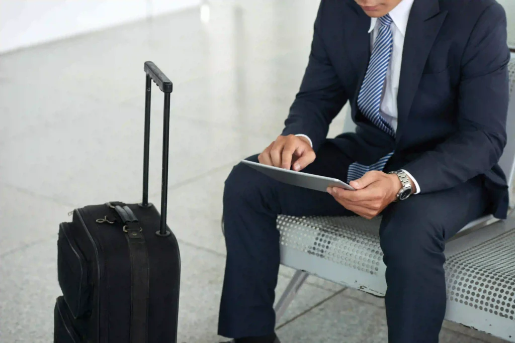 Persona seduta su una panchina in aeroporto, indossa un completo elegante e utilizza un tablet. Accanto, un trolley nero con manico esteso poggiato sul pavimento lucido.